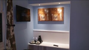 A room with a flat-screen TV mounted on the wall, a decorative wall piece, a small table with a coffee maker and cups, and a remote control on the table. - Ali Inn (Oxford)