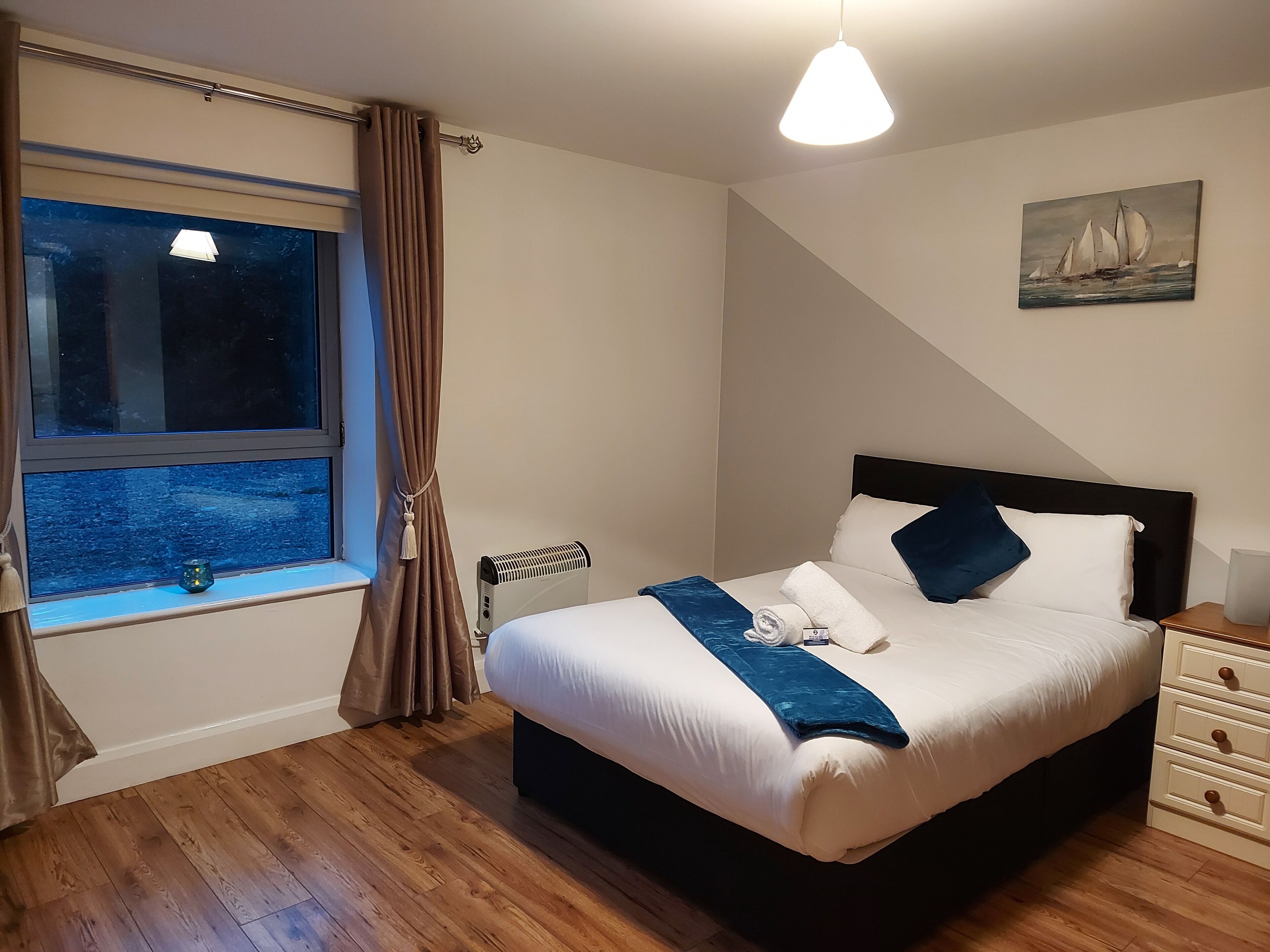 Una cama bien hecha con una decoración en blanco y azul, una ventana con cortinas, un radiador y un cuadro de veleros en la pared.