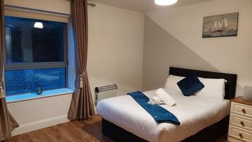 Una cama bien hecha con una decoración en blanco y azul, una ventana con cortinas, un radiador y un cuadro de veleros en la pared.