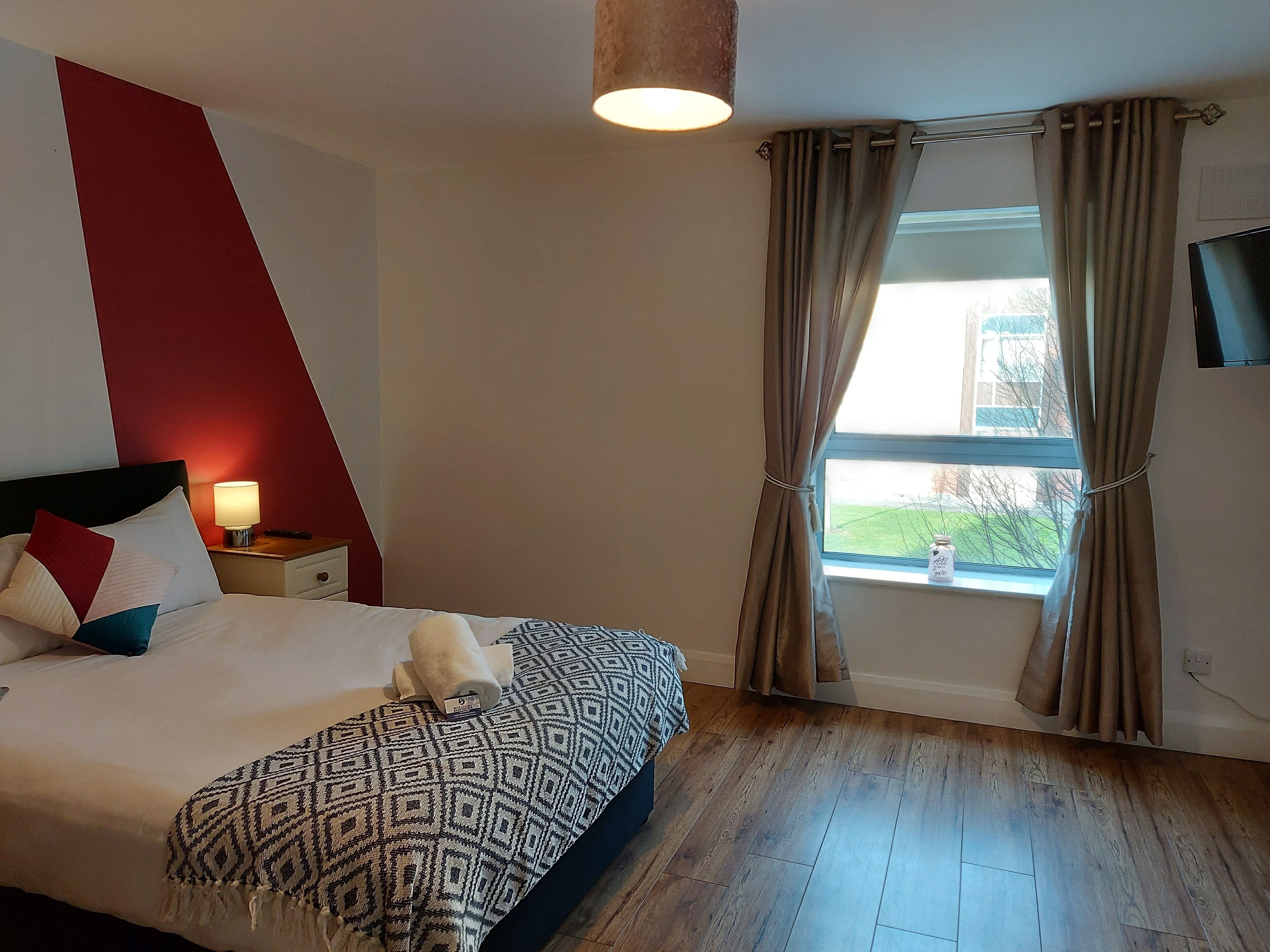 A hotel room with a bed, a nightstand, a lamp, a window with curtains, and a view of the outside.