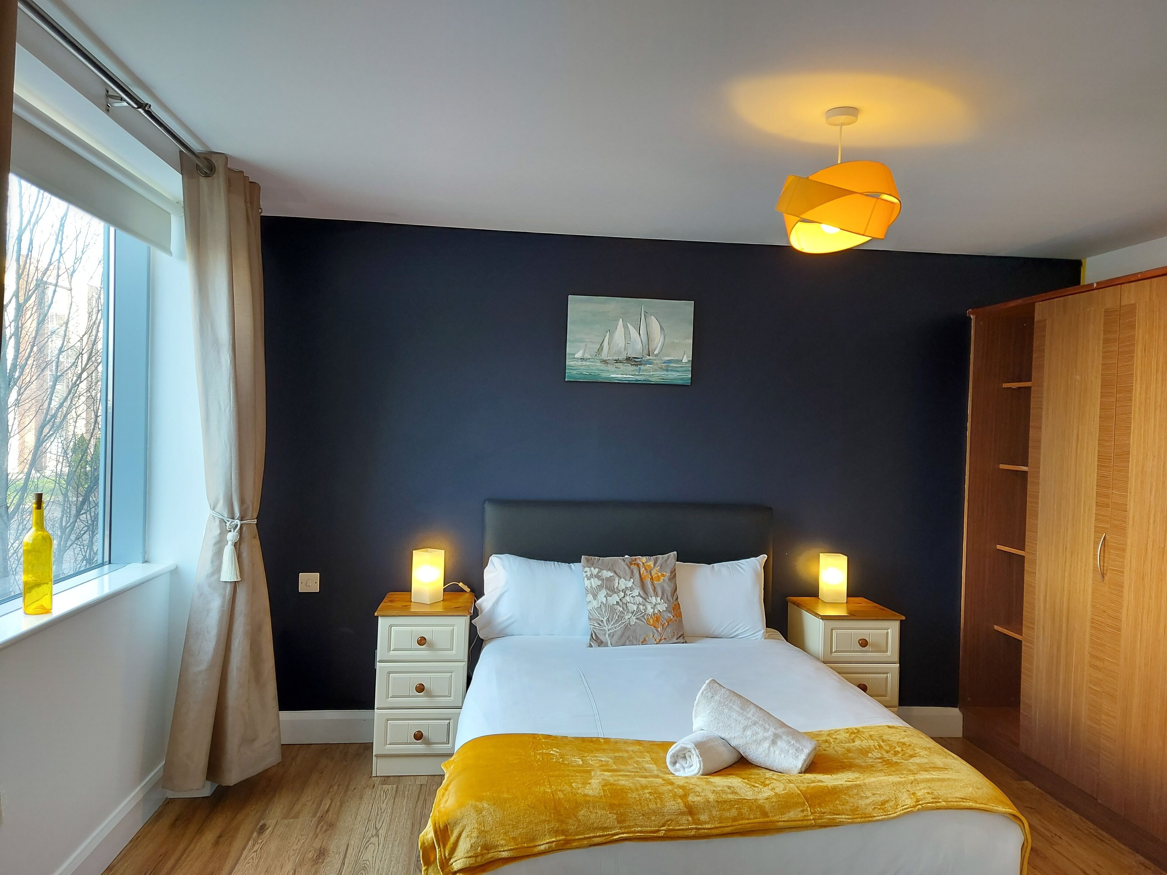 A bedroom with a bed, two bedside tables, a nightstand, a window with curtains, a painting, and a wardrobe.