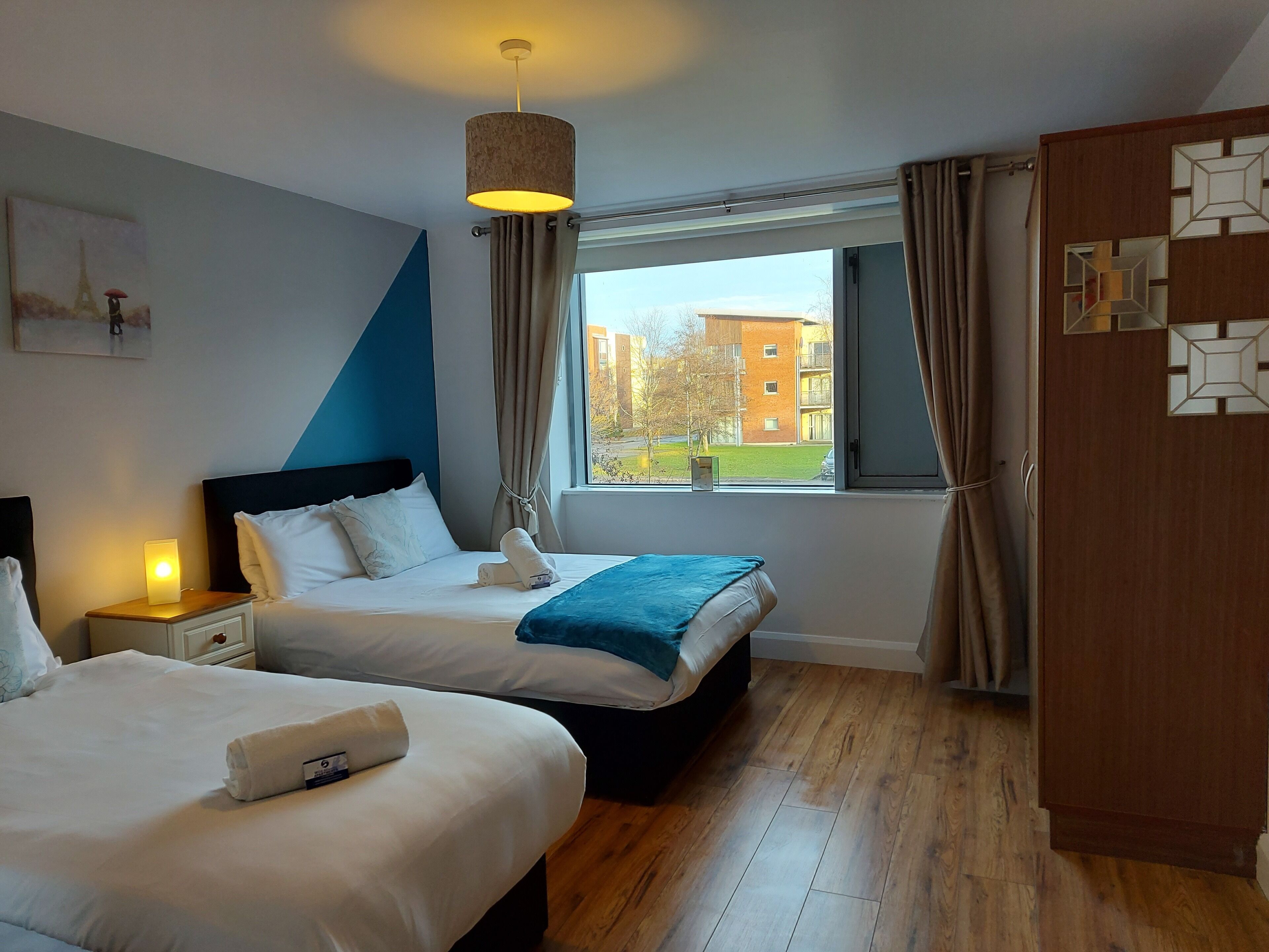A hotel room with two beds, a window with curtains, a wooden floor, and a painting of the Eiffel Tower.