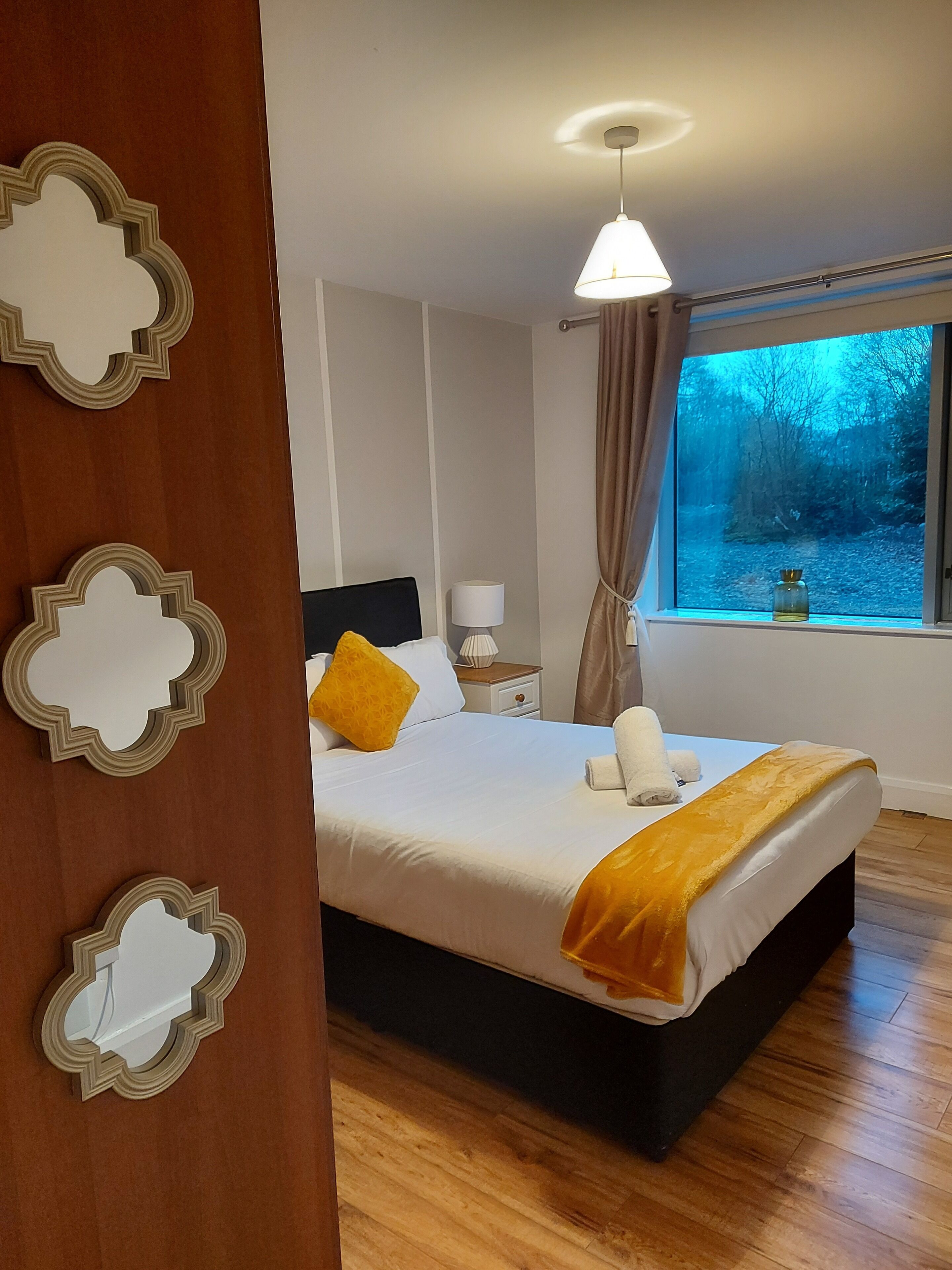 Une chambre à coucher avec un grand lit, une table de chevet avec une lampe, une fenêtre donnant sur des arbres et des miroirs décoratifs sur les murs.