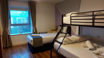 Quarto com beliches, janela com cortinas, mesa de cabeceira com abajur e vista para o exterior.