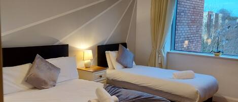 A hotel room with two beds, a nightstand with a lamp, a window with curtains, and a view of the outside.
