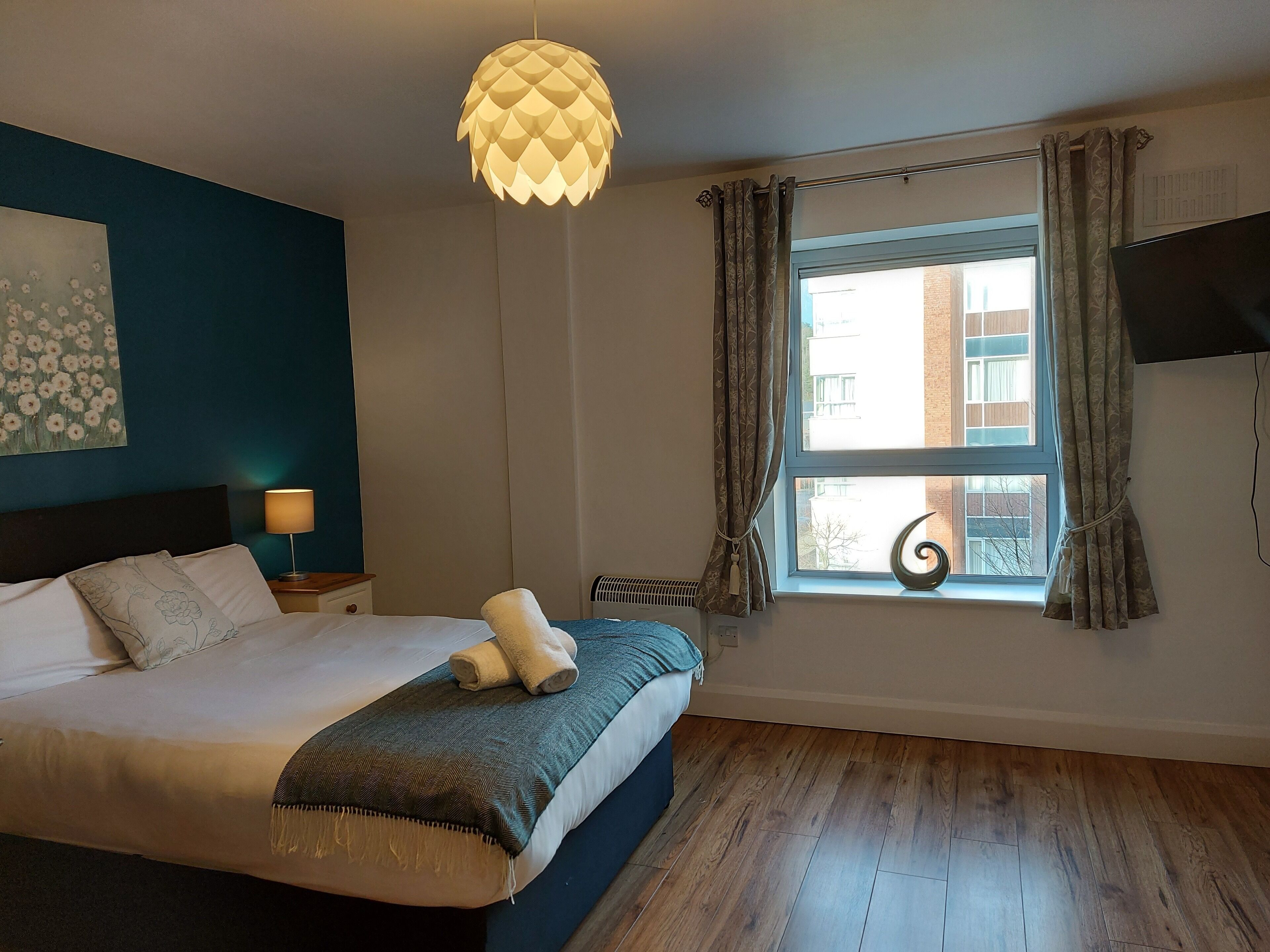 A bedroom with a bed, a bedside lamp, a window with curtains, a wall-mounted TV, and a ceiling light fixture.
