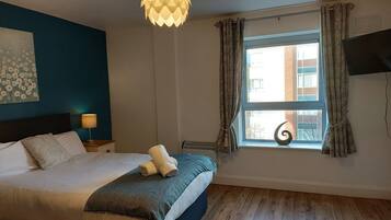 A bedroom with a bed, a bedside lamp, a window with curtains, a wall-mounted TV, and a ceiling light fixture.