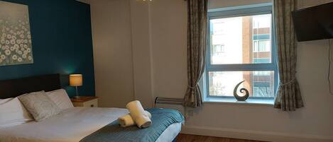 A bedroom with a bed, a bedside lamp, a window with curtains, a wall-mounted TV, and a ceiling light fixture.