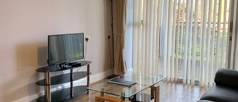 Ein modernes Wohnzimmer mit einem Glastisch, Ledersofas, einem Flachbildfernseher und einem großen Fenster mit vertikalen Jalousien.