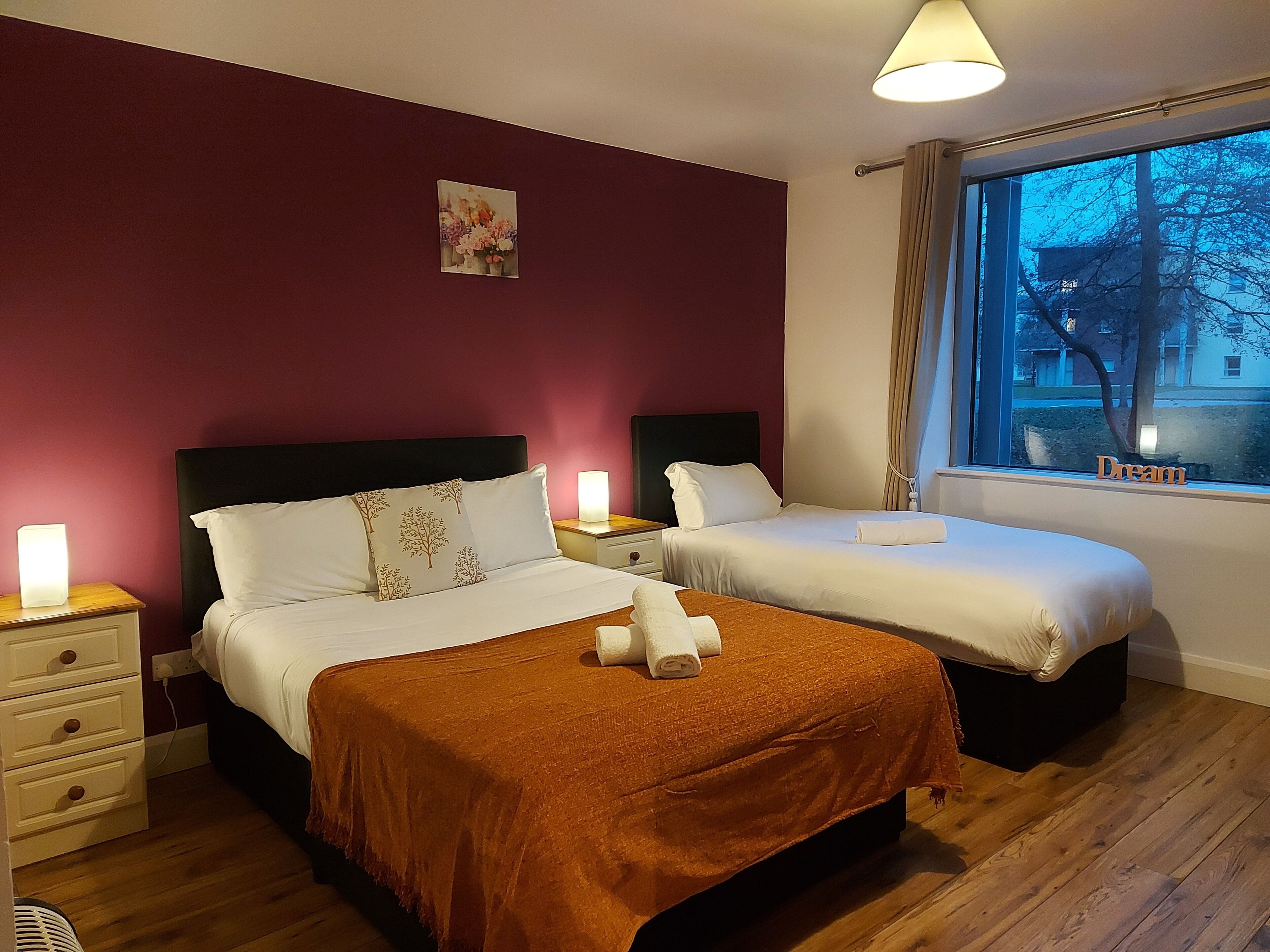 Een hotelkamer met twee bedden, een nachtkastje met een lamp, een dressoir en een raam met uitzicht op bomen en gebouwen.