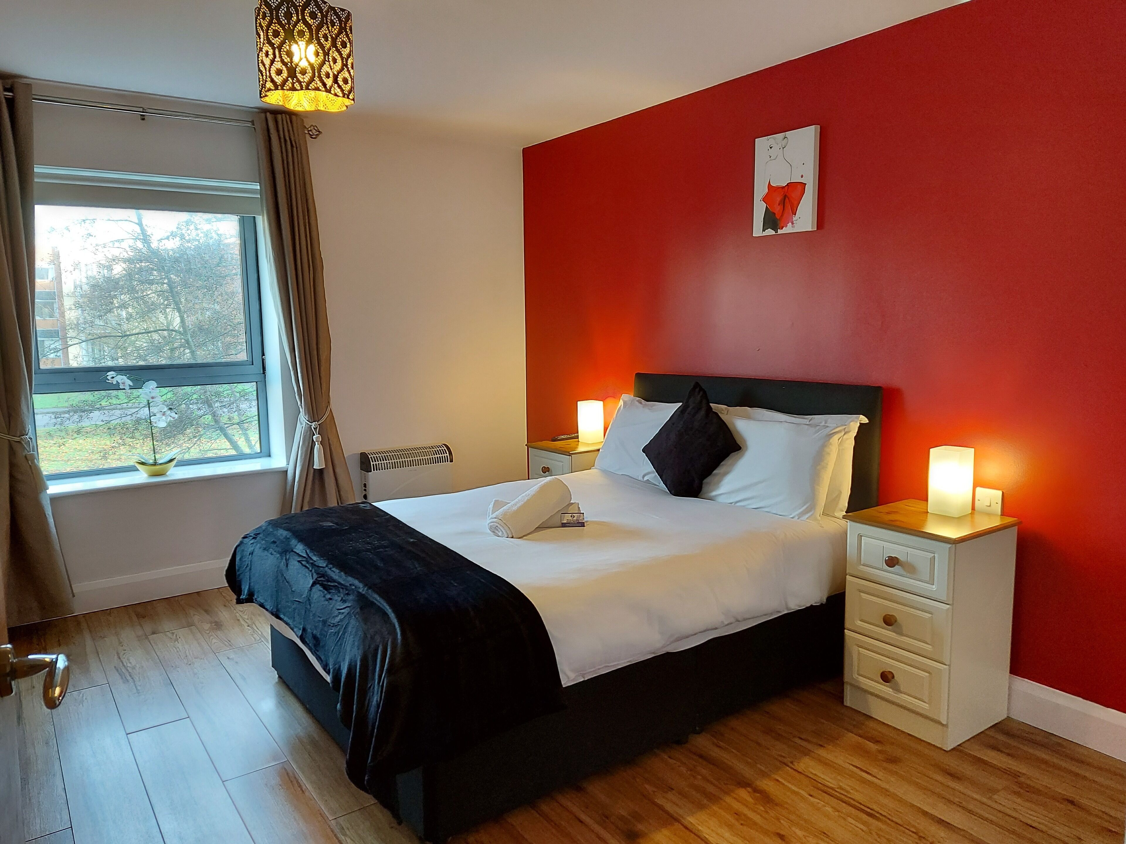 A bedroom with a bed, bedside tables, a window with curtains, a radiator, and a wall-mounted artwork.