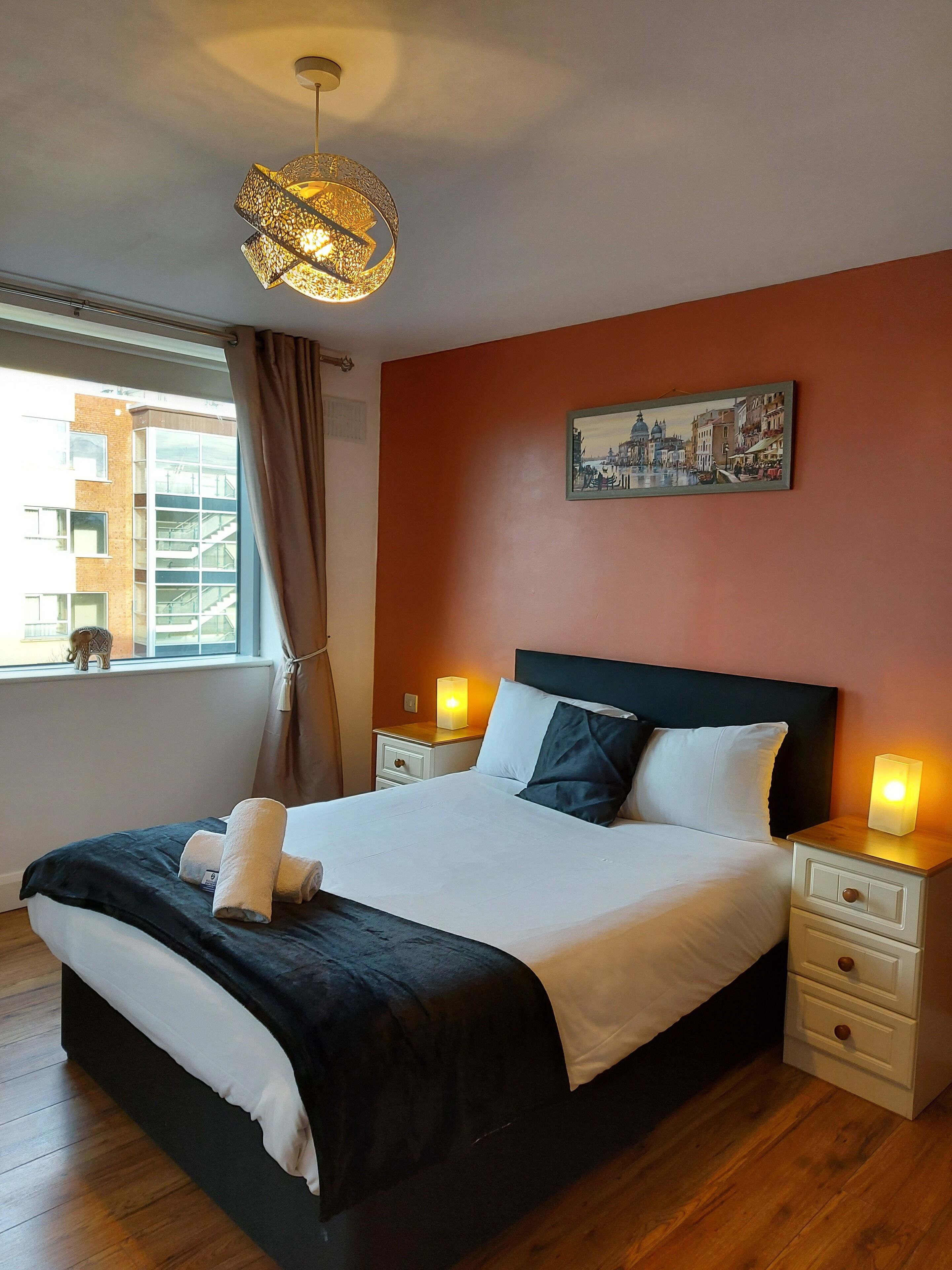 A neatly made bed with white and black bedding, two bedside tables with lamps, a painting on the wall, and a window with curtains.
