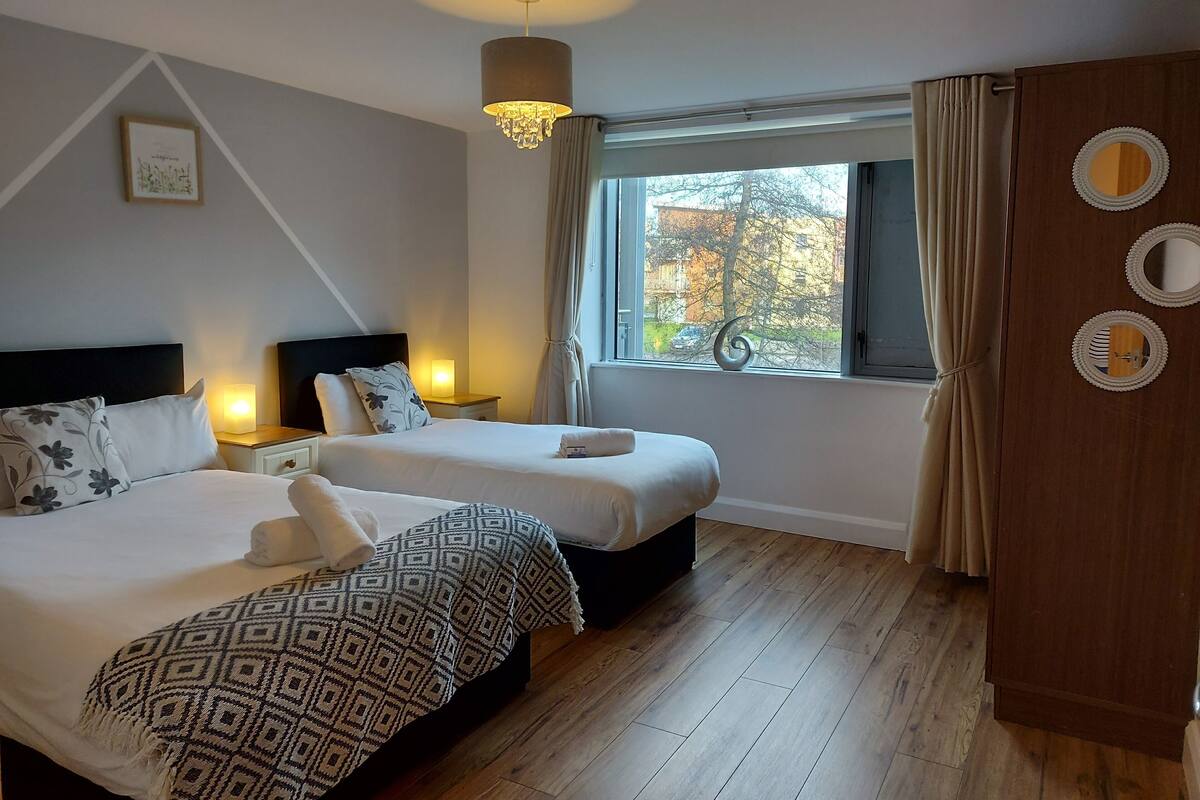 A hotel room with two beds, a large window with curtains, a wooden wardrobe, and a chandelier.