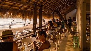 Un área de comedor al aire libre con mesas y sillas bajo un techo de paja, con vista a una playa.