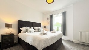 Ein modernes Schlafzimmer mit einem großen Bett, zwei Nachttischen, einem Schränkchen, einem Fenster mit Vorhängen und einer Heizung.