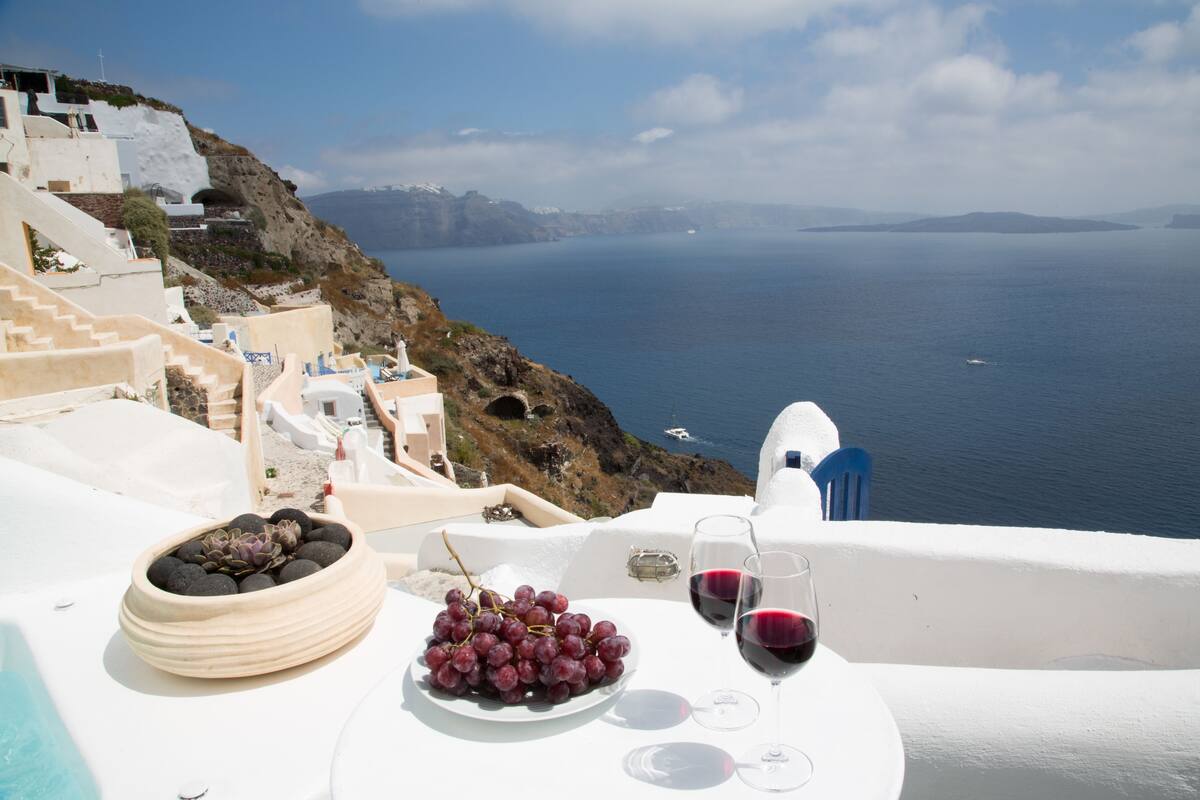 A table with a bowl of grapes and wine glasses, overlooking a coastal view with white buildings and a clear sky.