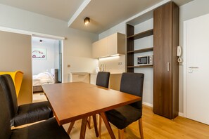 A modern living room with a wooden dining table, black chairs, a built-in bookshelf, a kitchen area with a microwave, and a white door.