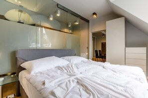 A modern bedroom with a large bed, a glass partition wall, and a view of a dining area. - Apartments Bohemia Rhapsody (Karlovy Vary)