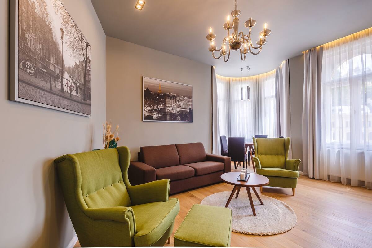 Ein Wohnzimmer mit einem braunen Sofa, grünen Sesseln, einem runden Couchtisch und einem Kronleuchter.