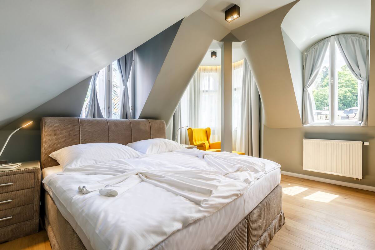 Ein Schlafzimmer mit einem großen Bett, einem Nachttisch, einem Sessel und einem Fenster mit Vorhängen.