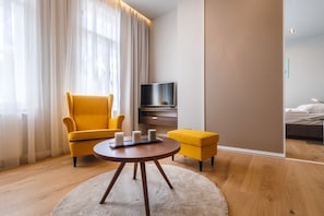 A modern hotel room with a yellow armchair, a round wooden coffee table with candles, a flat-screen TV, and a bed with white linens.