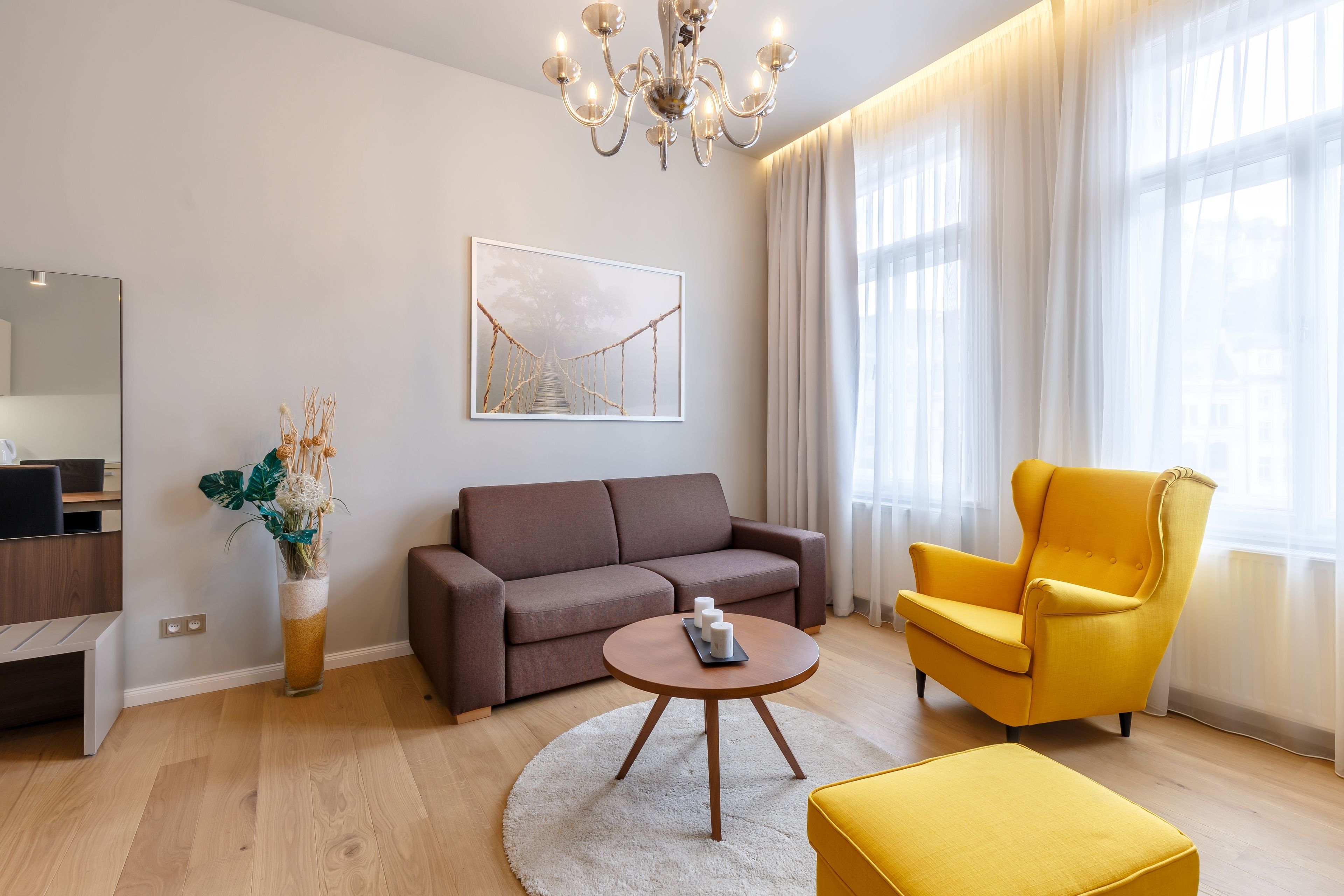 Un salon moderne avec un canapé marron, un fauteuil jaune, une table basse ronde et une grande fenêtre ornée de rideaux légers.