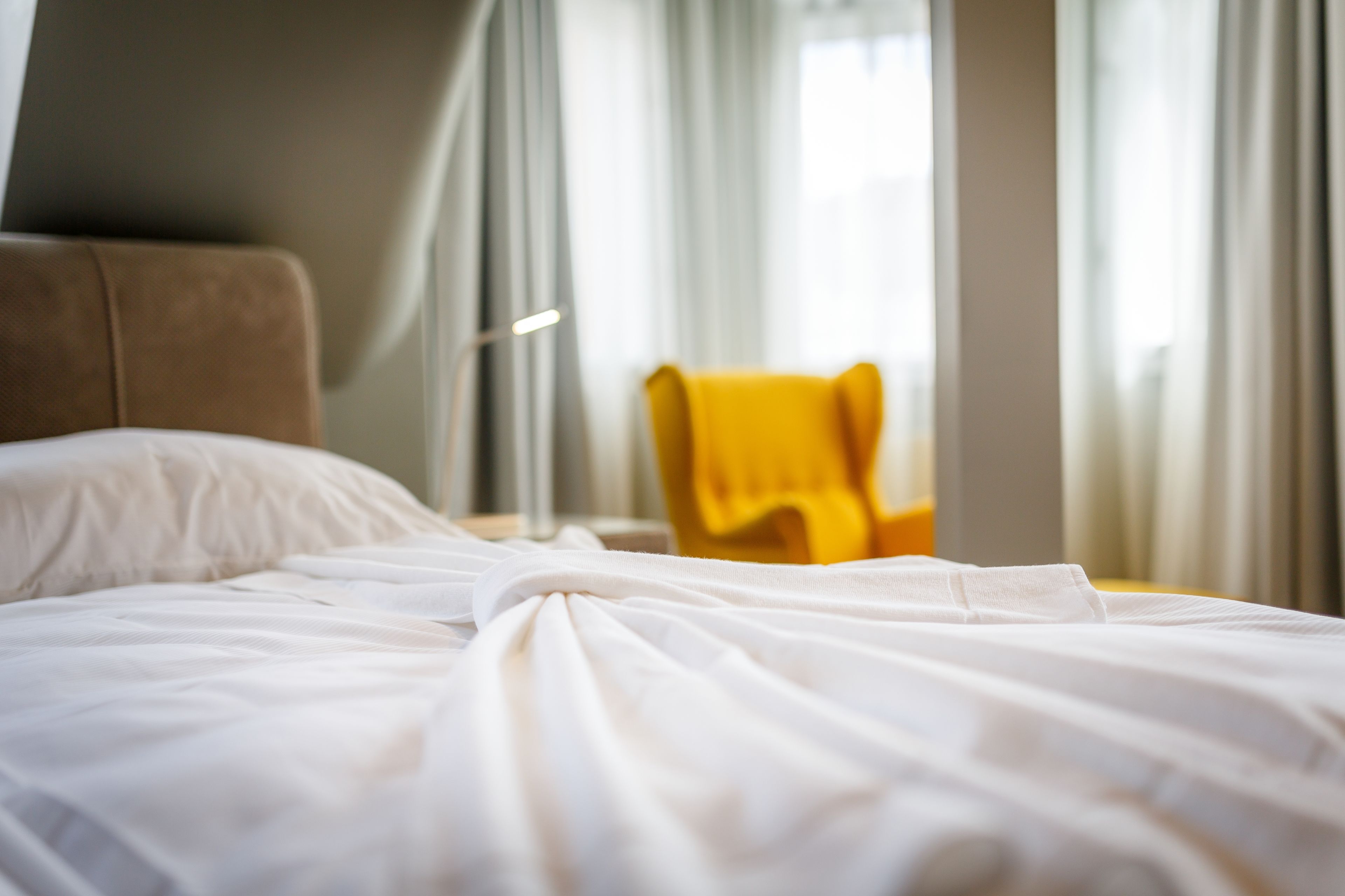 A hotel room with a bed, a yellow chair, and a window with curtains.