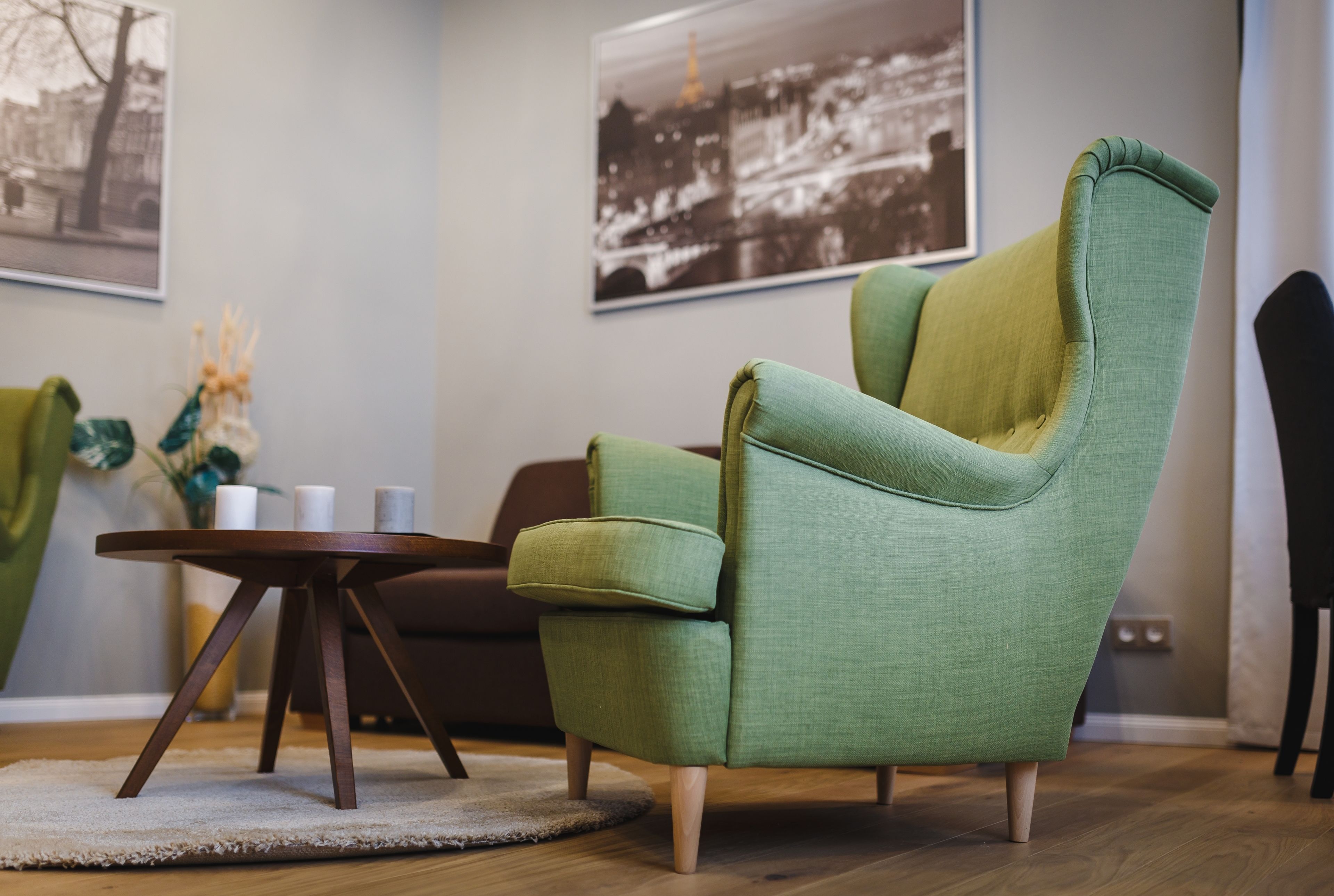 Un fauteuil vert aux pieds en bois, une table ronde en bois avec deux bougies, et un tableau représentant un paysage urbain, encadré, accroché au mur.