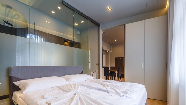 Quarto moderno com uma cama grande, um painel de vidro e uma área de refeições visível através do painel.