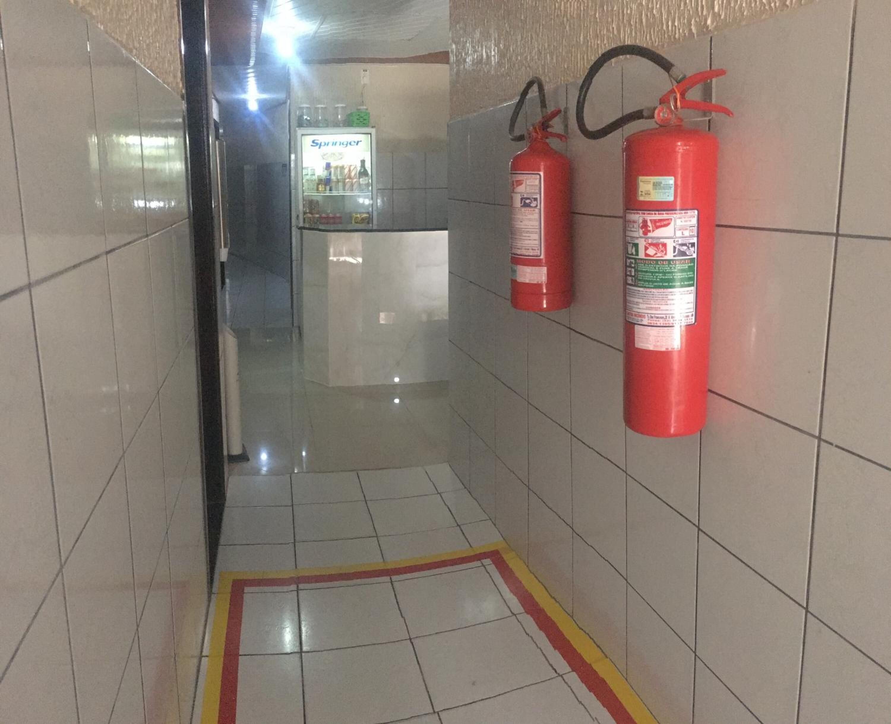 Un pasillo con paredes y suelo de baldosas, dos extintores rojos montados en la pared y una máquina expendedora con el rótulo 'Springer' al fondo.