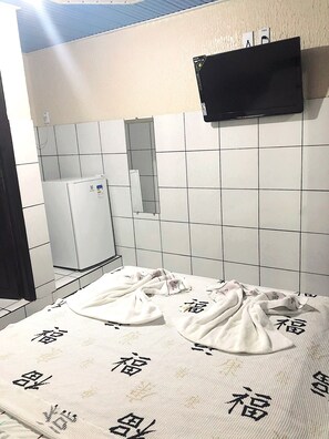 Een badkamer met witte betegelde muren, een wit wastafelmeubel en een spiegel. Er hangt een zwarte televisie aan de muur en er ligt een handdoek met Japanse karakters op de vloer.