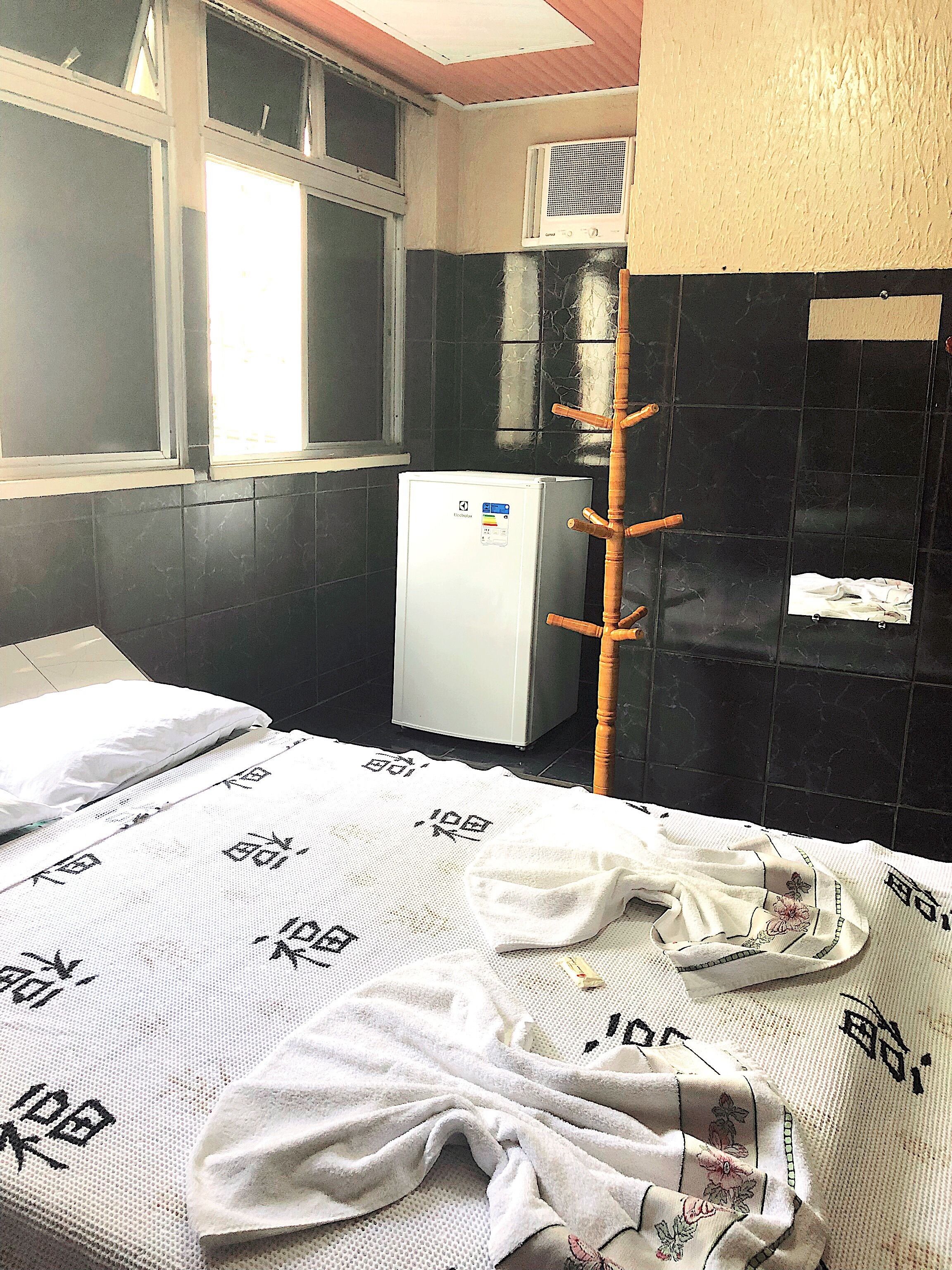 Una habitación pequeña con una cama, un refrigerador blanco, un ventanal y un aire acondicionado de pared.