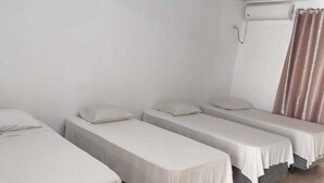 Una habitación con tres camas individuales, cada una con colchón y almohada blancos. Tiene una ventana con cortinas y un equipo de aire acondicionado instalado en la pared.