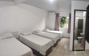 Una habitación con tres camas individuales, una mesita, una ventana con cortinas y un baño visible a través de una puerta abierta.