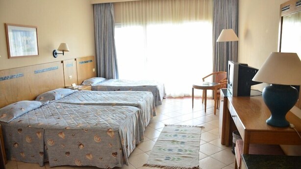 A hotel room with two single beds, a desk with a lamp, a television, and a small table. - Cyrene Island Hotel (sharm el sheikh)