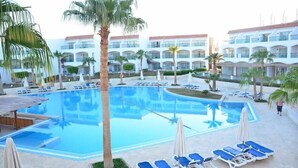 A hotel pool area with multiple lounge chairs and umbrellas, surrounded by palm trees and a building with balconies. - Cyrene Island Hotel (sharm el sheikh)