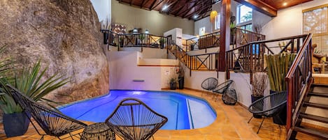 An indoor swimming pool with a rock feature, surrounded by wicker chairs, a staircase, and a wooden ceiling.