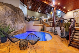 Una piscina interior con una formación rocosa, rodeada de sillas de mimbre, una escalera y un techo de madera.