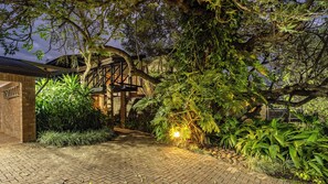 Un chemin de briques mène à un escalier en bois, lui-même entouré d’une végétation luxuriante, et aboutit à un bâtiment en briques.