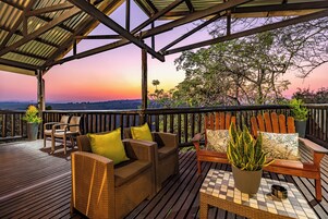 Una terraza de madera con mobiliario de mimbre y plantas en macetas, con vistas a un paisaje pintoresco.