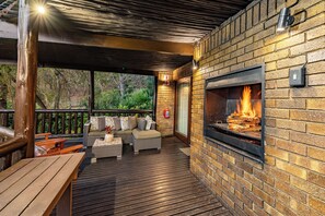Een terras buiten met een bakstenen open haard, een houten terras en een zithoek met kussens.