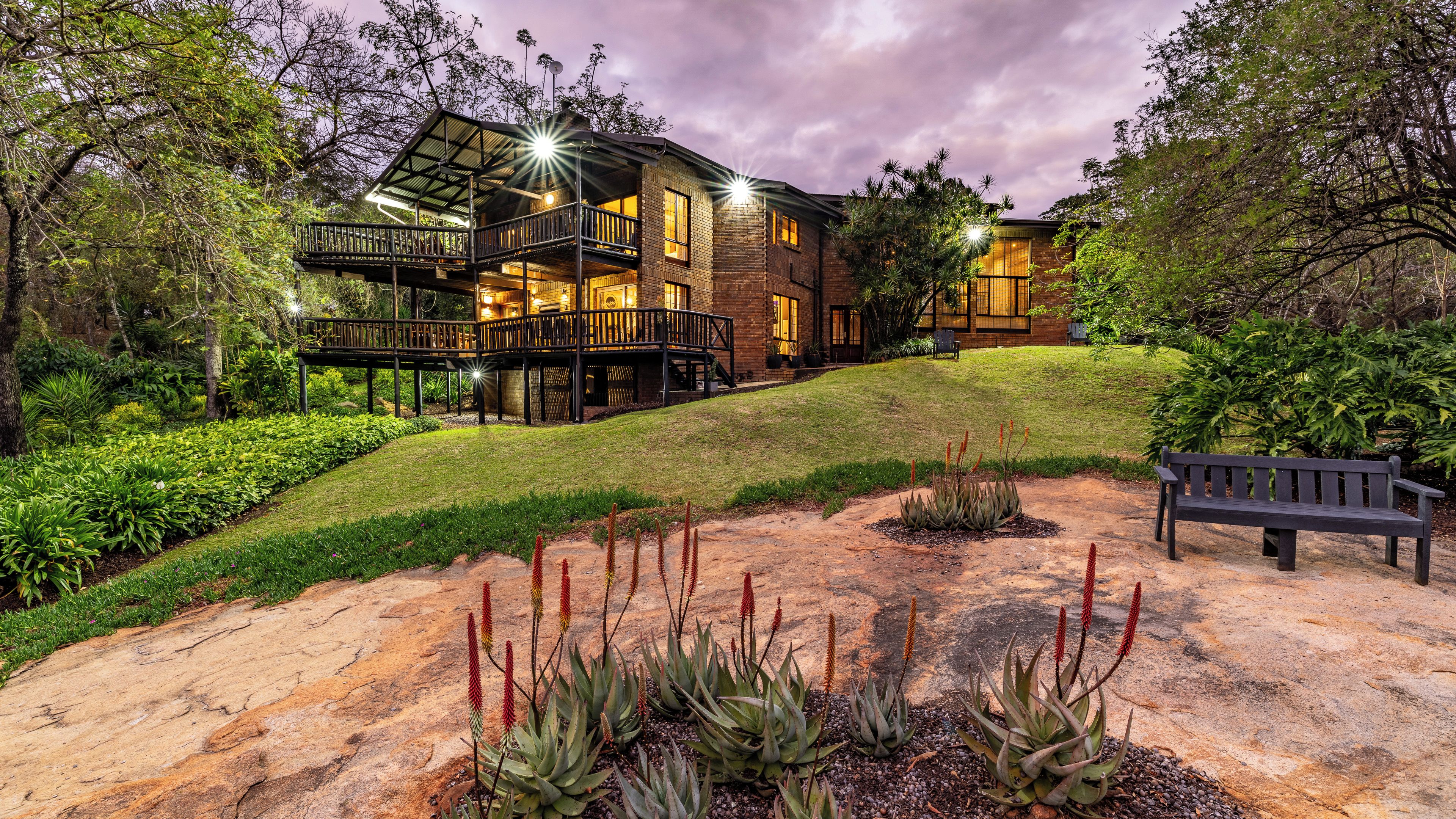A two-story building with a balcony, surrounded by a landscaped garden with various plants and a bench.