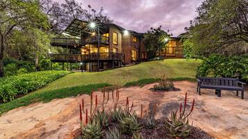 A two-story building with a balcony, surrounded by a landscaped garden with various plants and a bench.
