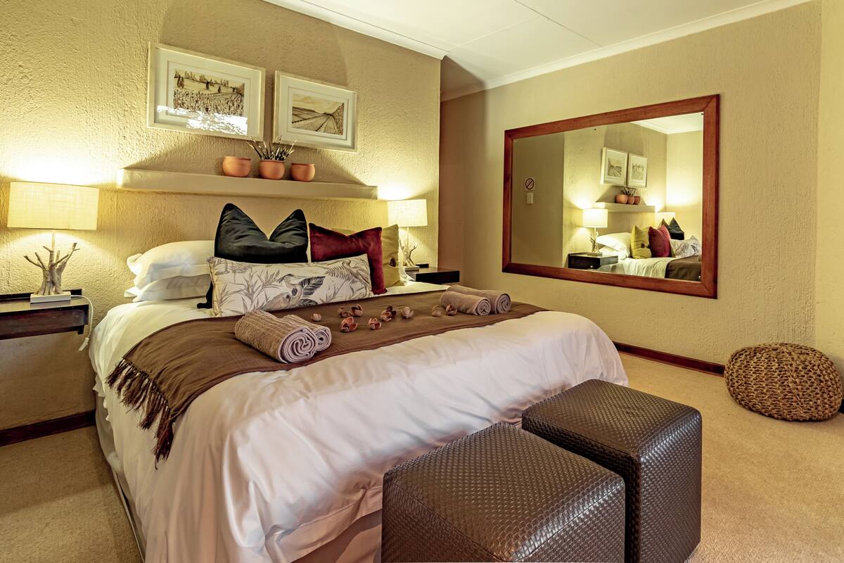 A neatly made bed with pillows, a bedside table with a lamp, a large mirror, and a small round seating area.
