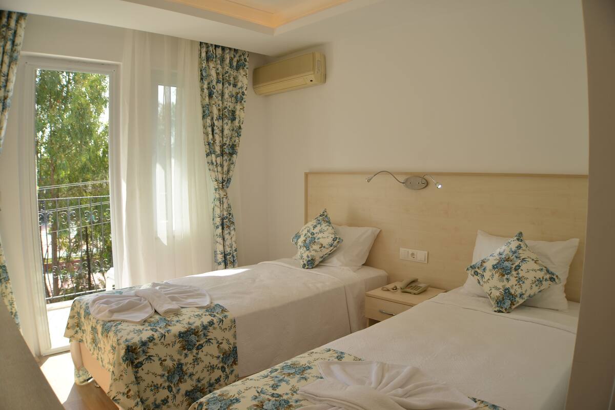 A hotel room with two beds, a window with curtains, an air conditioning unit, and a bedside table with a phone.