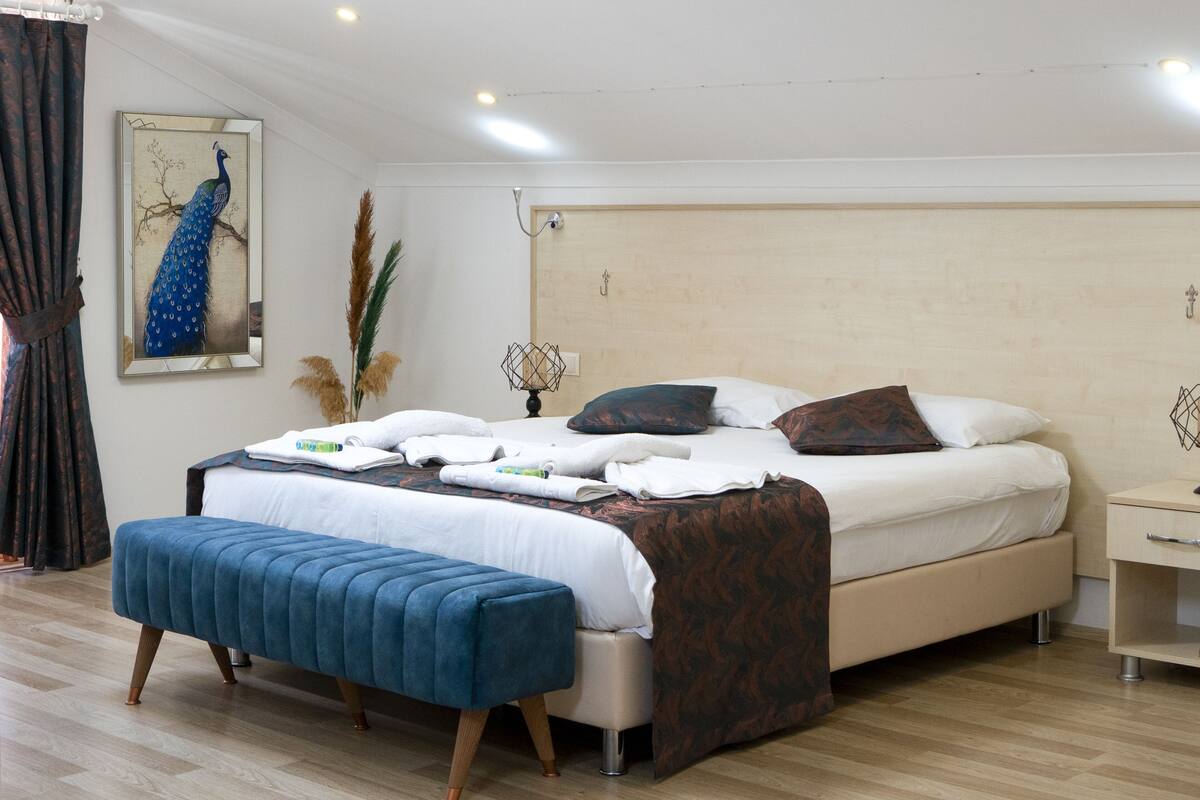 A modern bedroom with a large bed, a blue bench, and a peacock painting on the wall.