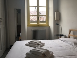 A hotel room with a bed, a window with a view of a tiled roof, a radiator, and a bedside table with a lamp. - Gioia 11 (Turin)