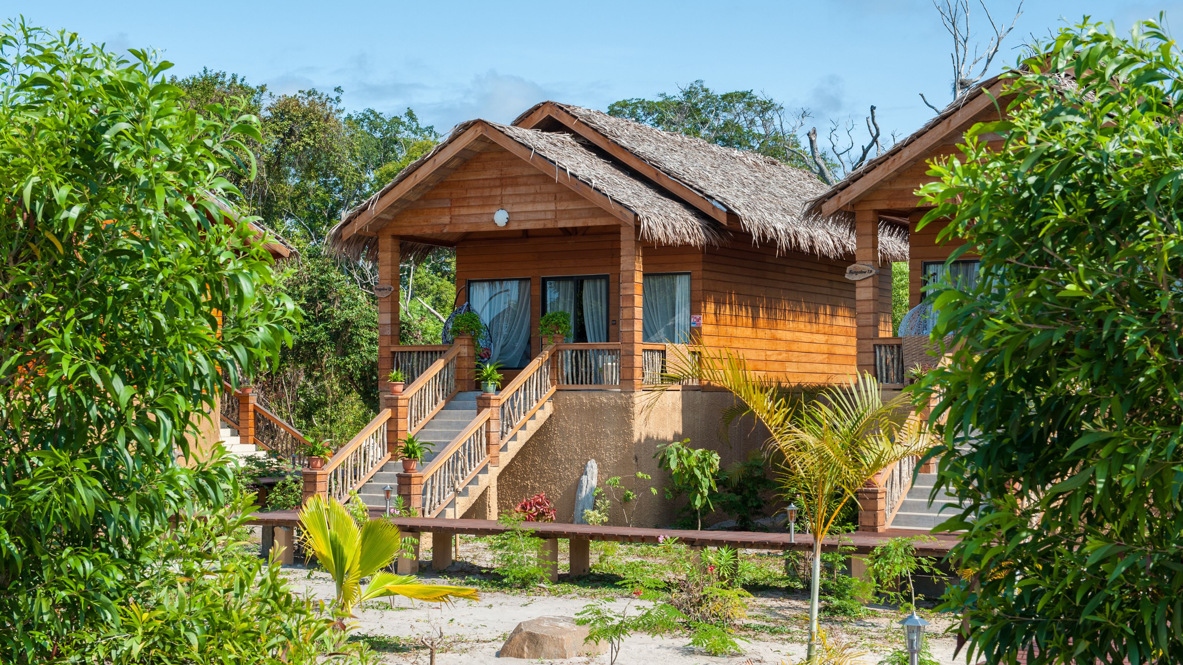A wooden bungalow with a thatched roof, a covered porch, and a staircase leading to the entrance.