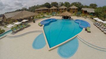 A large swimming pool with multiple lanes, surrounded by lounge chairs and umbrellas, with thatched-roof buildings in the background.