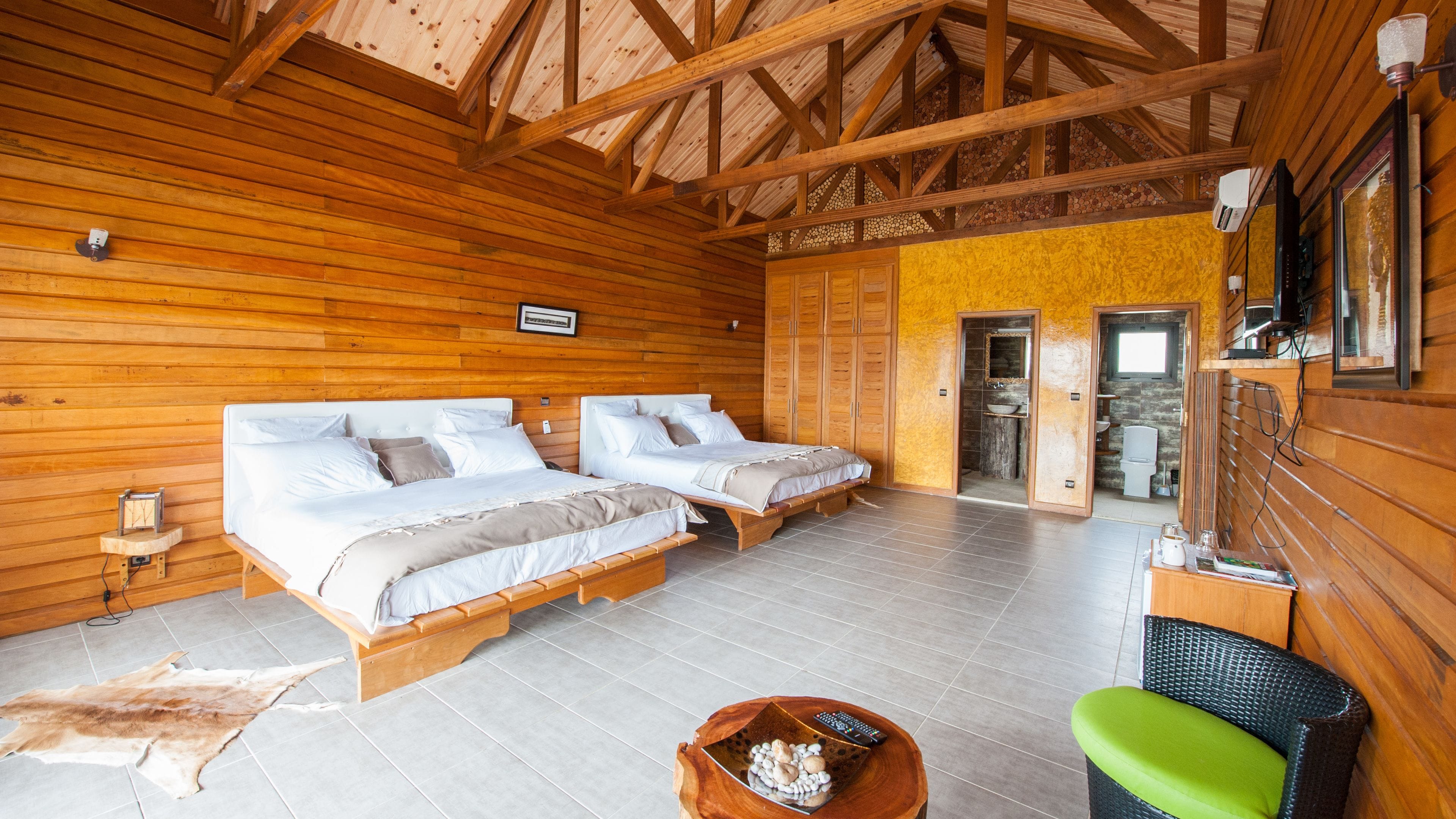 A wooden cabin with two beds, a small table, a green chair, and a bathroom visible through an open door.